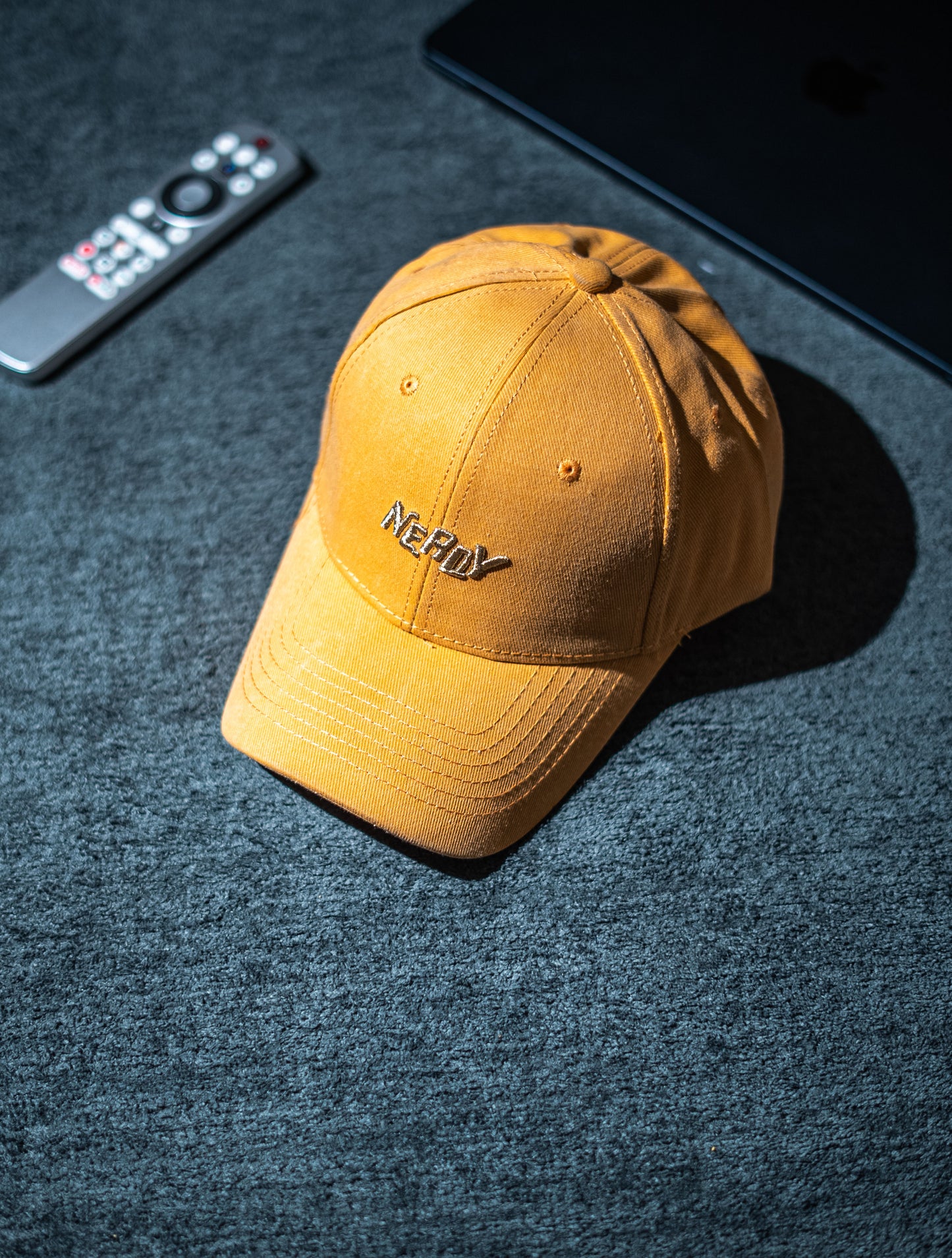 Yellow cap with a brand logo on a dark textured surface next to a remote control and smartphone in Pakistan by Caps Nation