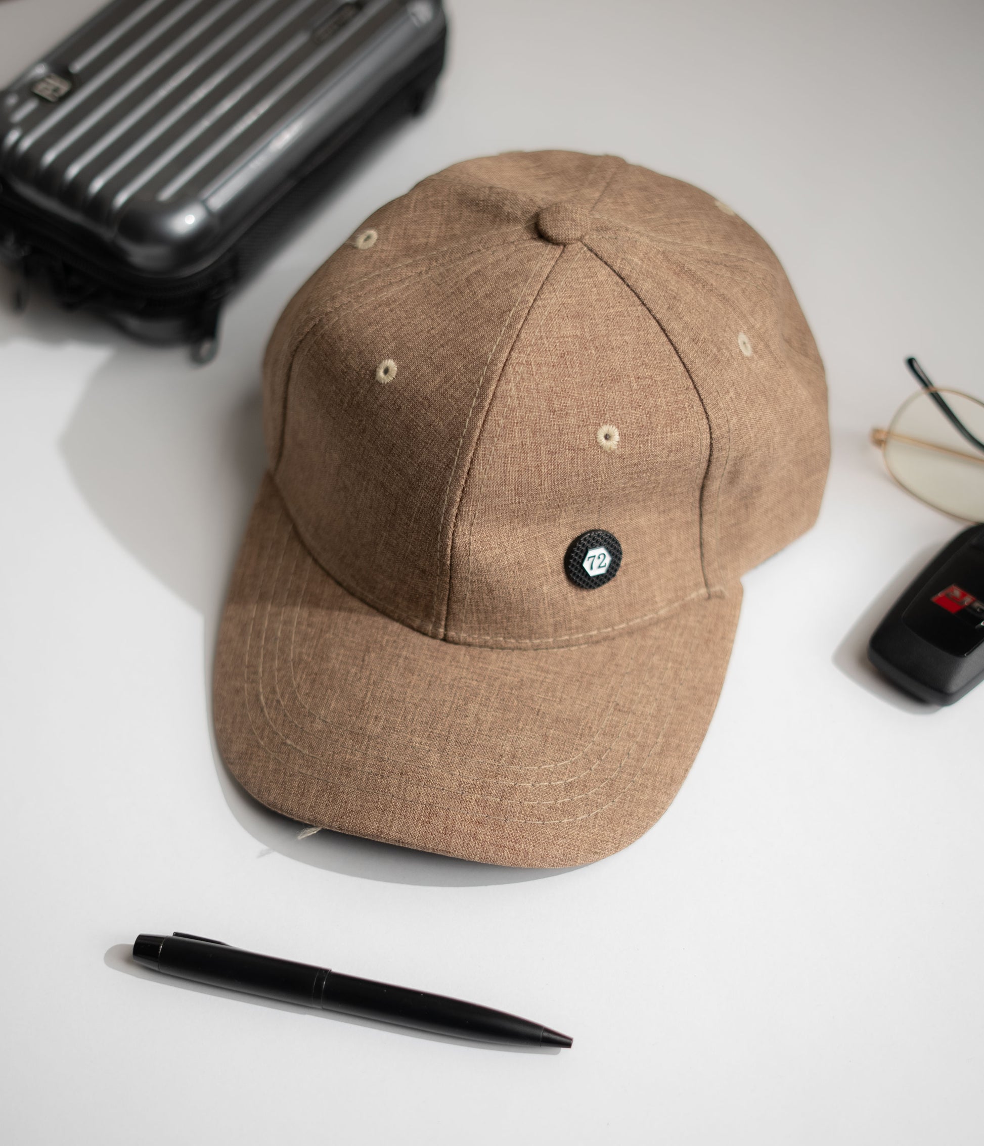 Brown cap with a small logo on a white surface with a suitcase, pen, and glasses in Pakistan by Caps Nation