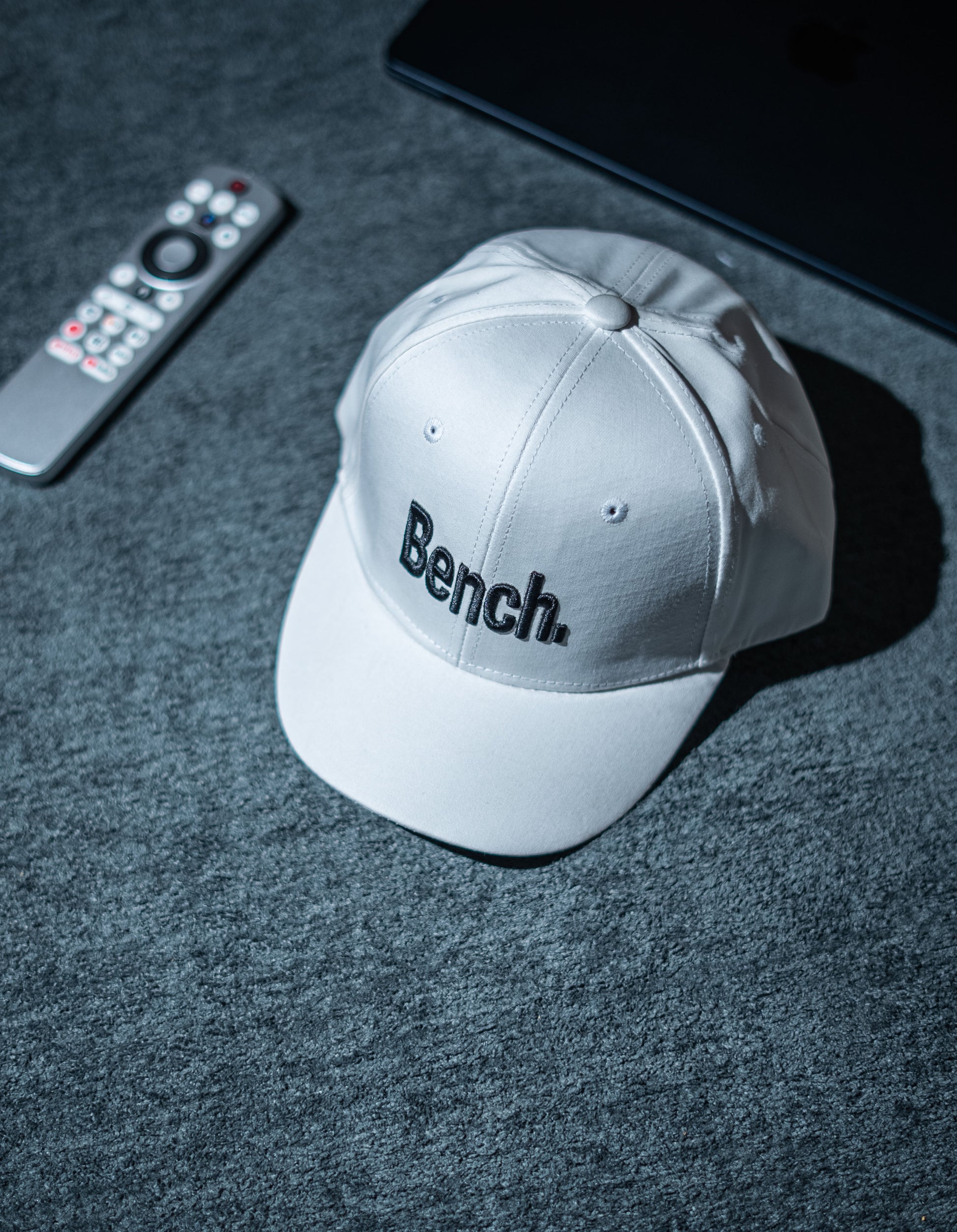 Gray cap with 'Bench' logo on a dark textured surface with remote control and tablet in Pakistan by Caps Nation