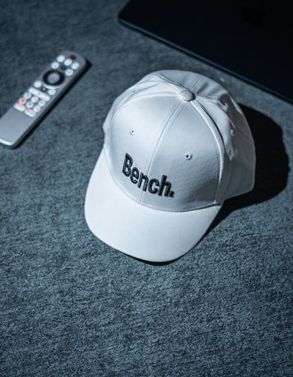 Gray cap with 'Bench' logo on a dark textured surface with remote control and tablet in Pakistan by Caps Nation