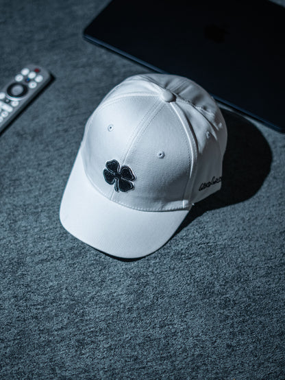 White cap with a black four-leaf clover emblem on a dark surface in Pakistan by Caps Nation