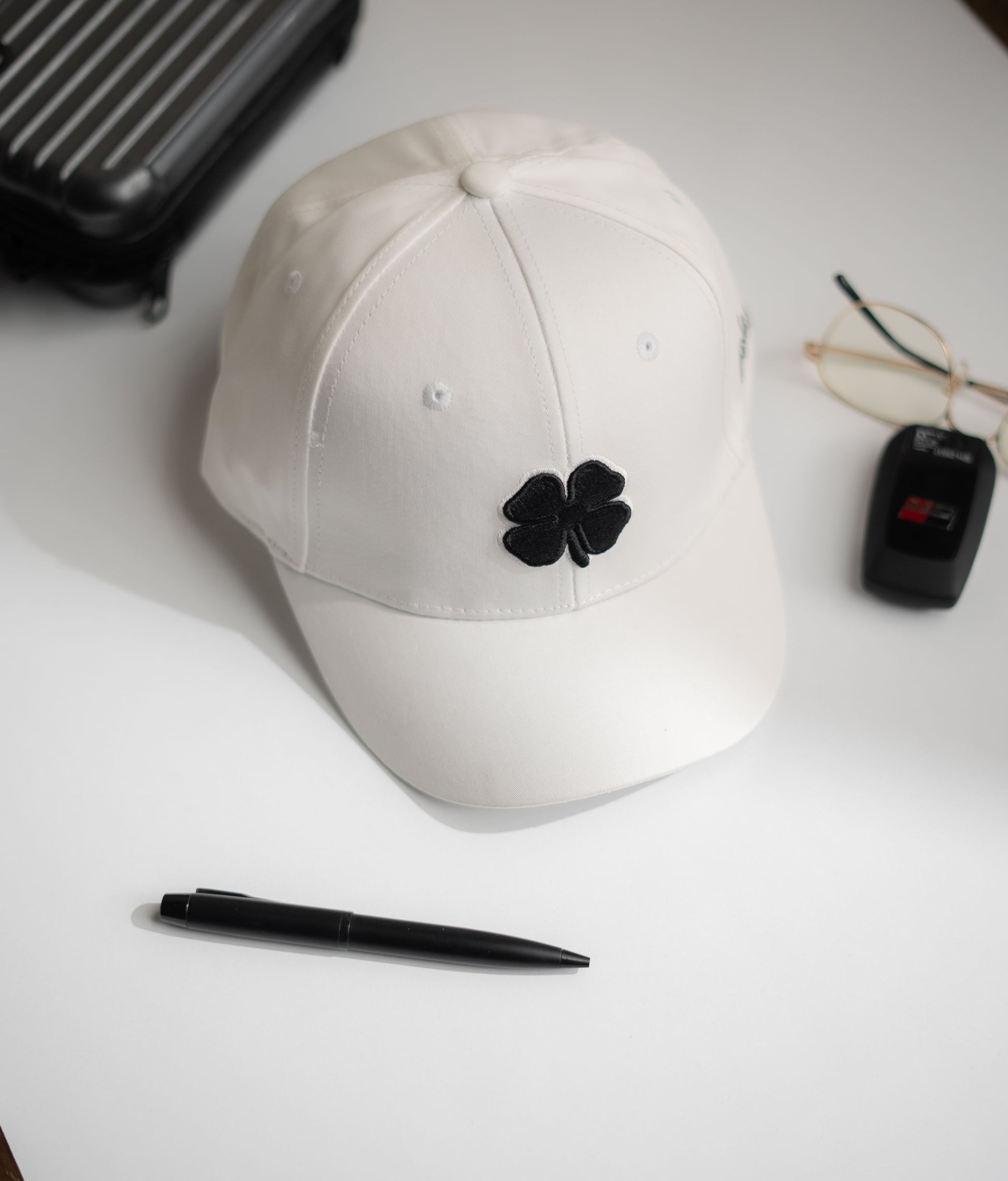 White cap with black flower emblem on a white surface with a pen and small black device in Pakistan by Caps Nation