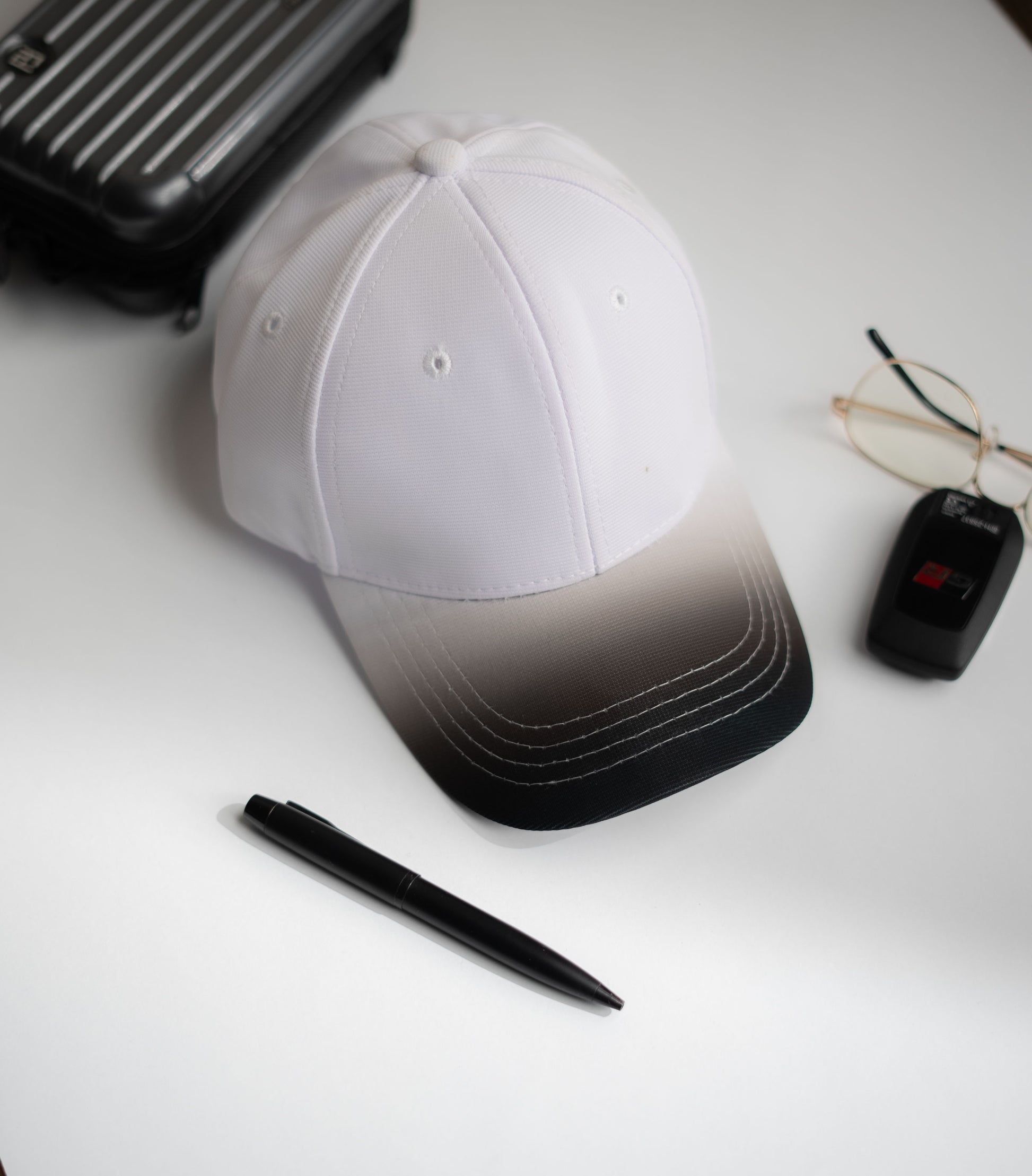 White cap with black gradient, pen, glasses, and suitcase on a white surface in Pakistan by Caps Nation