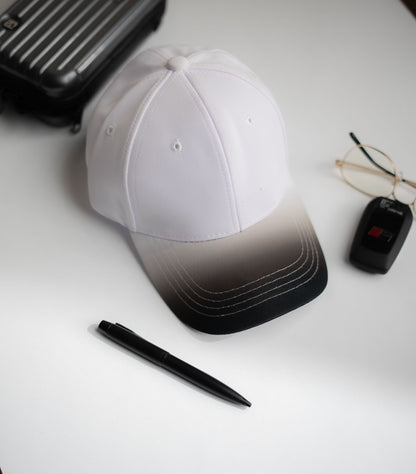 White cap with black gradient, pen, glasses, and suitcase on a white surface in Pakistan by Caps Nation