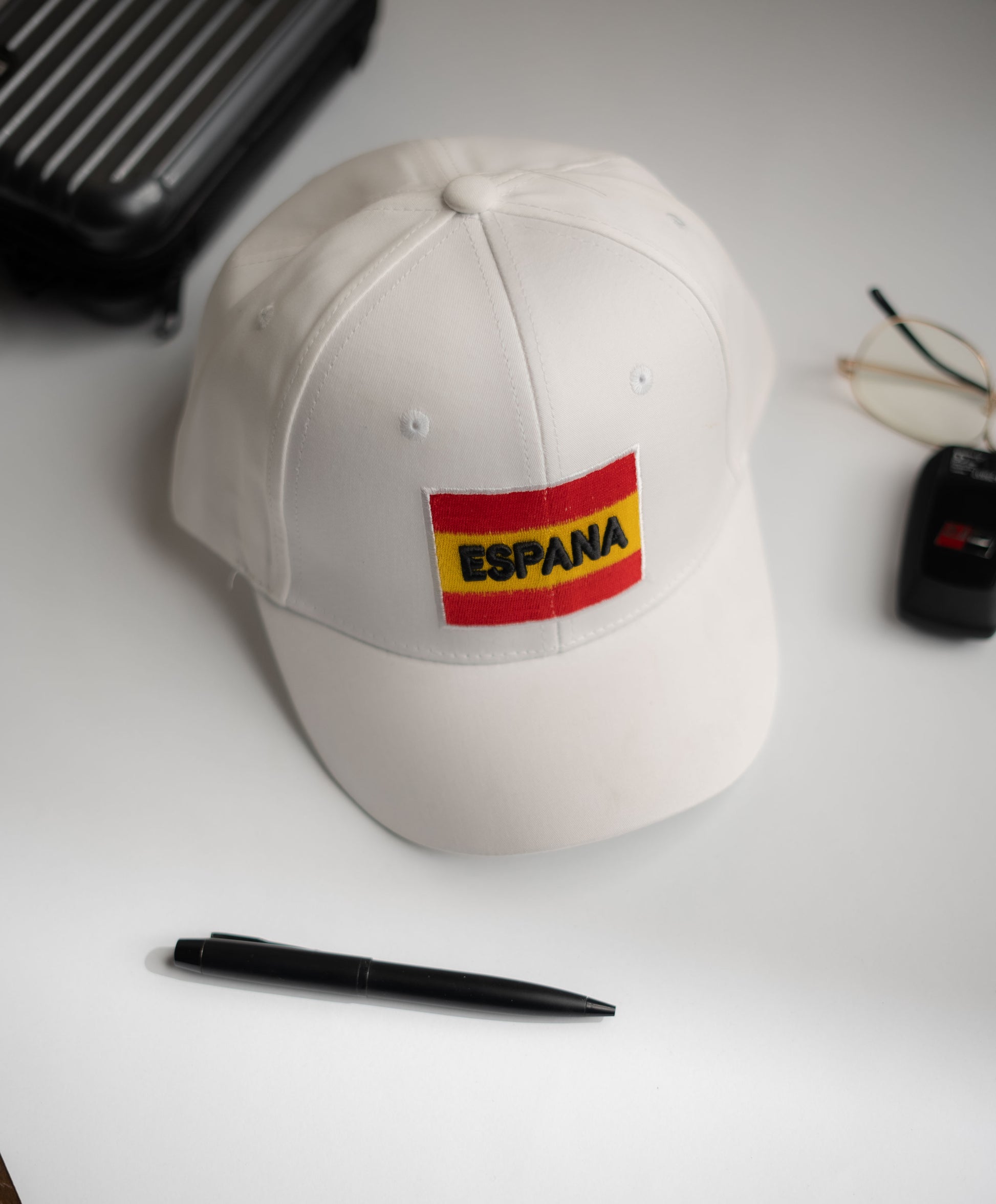 White cap with 'ESPAÑA' patch on a white surface with a pen and glasses in Pakistan by Caps Nation