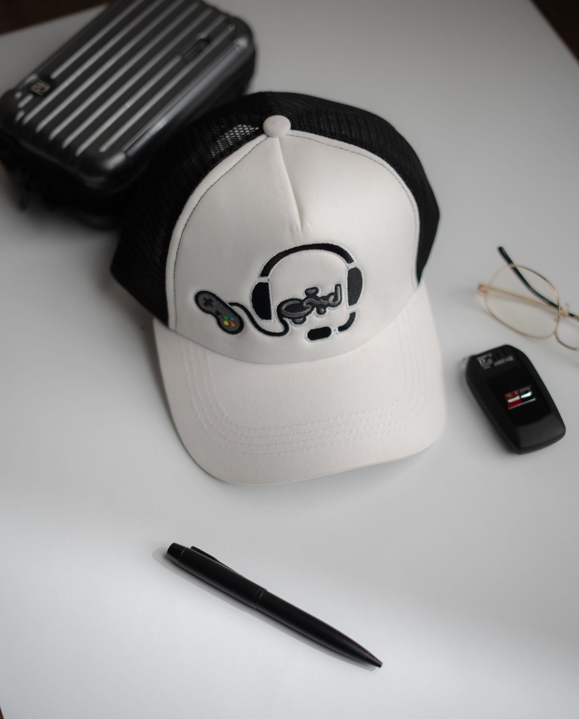 White cap with black logo on a white surface with a pen, glasses, and black case in Pakistan by Caps Nation